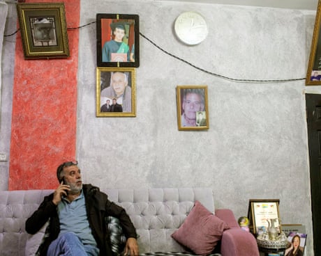 A man on a sofa talks on a phone with family portraits and a clock on the wall behind him