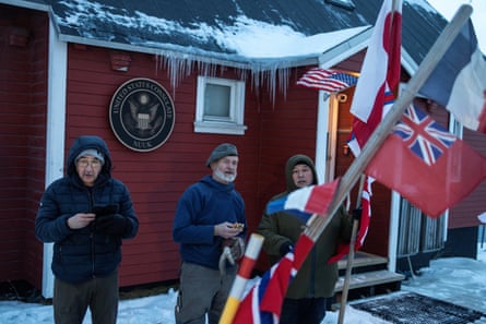 Three people with flags, including Greenland’s outside the single-storey US consulate. Snow lies on the ground and on part of the building’s roof.