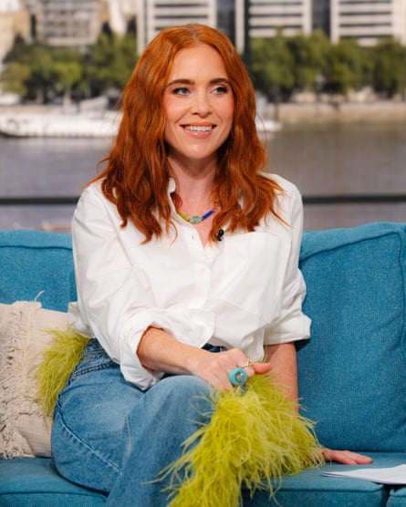 she is in her early 40s and has long red hair. She wears a white linen shirt and blue jeans.