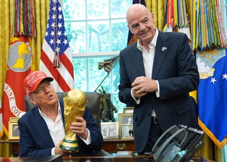 Trump seated holding World Cup trophy with. US flag behind and Infantino standing