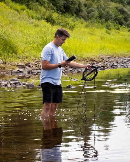 A man in shorts and T-shirt stands in a river holding a wire and electronic device
