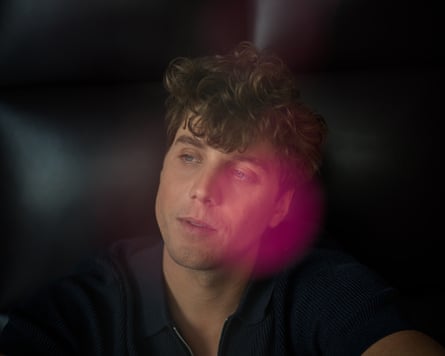 A photograph of Lukas Gage, looking to the side. There is a pink bokeh effect on the side of his face in the picture.
