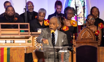Sharpton speaks into a microphone. Behind him are a group of people with sombre expressions