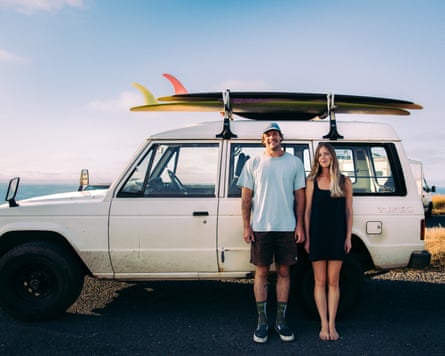 The pair stand beside a van with two surfboards on a roof rack, smiling, as the sun goes down over the sea