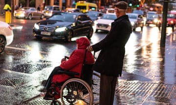 An older man waits at a pedestrian crossing pushing a person in a wheelchair