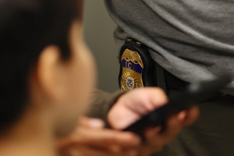 A blurry child's head in the foreground, with adult hands operating a cellphone and an ICE badge on a waistband beyond.