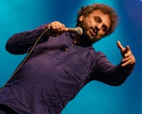 Nish Kumar leans back, gesturing with one hand while holding a microphone on stage
