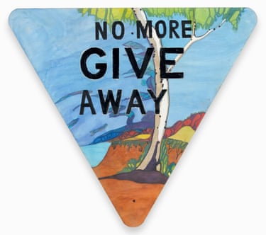 A give way road sign that has been painted so that it says ‘no more give away’ and has a gum tree on a hill by the coast