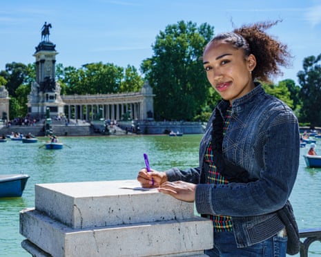 A woman stands in front of a lake smiling at the camera as she puts pen to paper.