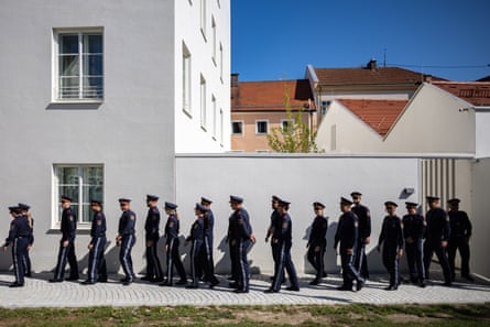 A line of officers in dark uniforms walk in front of the white-walled building