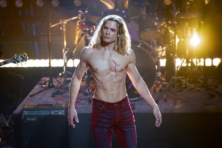 A bare-chested man with blond hair with streaks of blood on his chest, weaering jeans, with a drum kit behind him