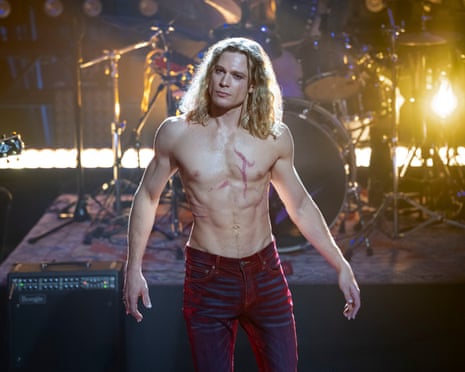 A young bare chested and ripped young man with leather trousers and long fair hair wearing makeup