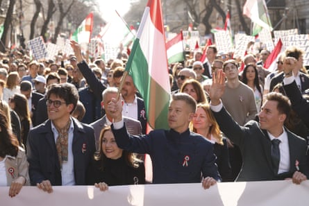 Magyar, front centre, during a rally in Budapest. People hold flags behind him with some raising their hands in the air. It is sunny in a wide, tree-lined street and the people around Magyar look happy and smiling.