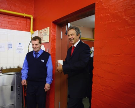 Blair walks into a stark room, built of bricks painted red, with a mug of tea, grinning