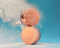 A round loose powder compact stacked upright on top of another one as powder bursts around them against a blue background
