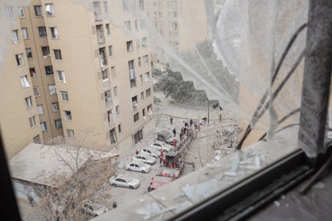 View from out of a shattered window looking down on dust and debris below. Apartment blocks opposite have blown-out windows
