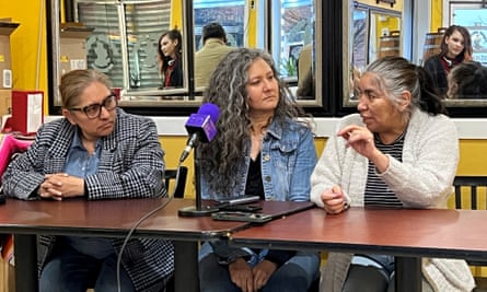 At a long table sit three Latina women, with a purple-covered microphone in the middle. The woman on the right speaks as the others listen.