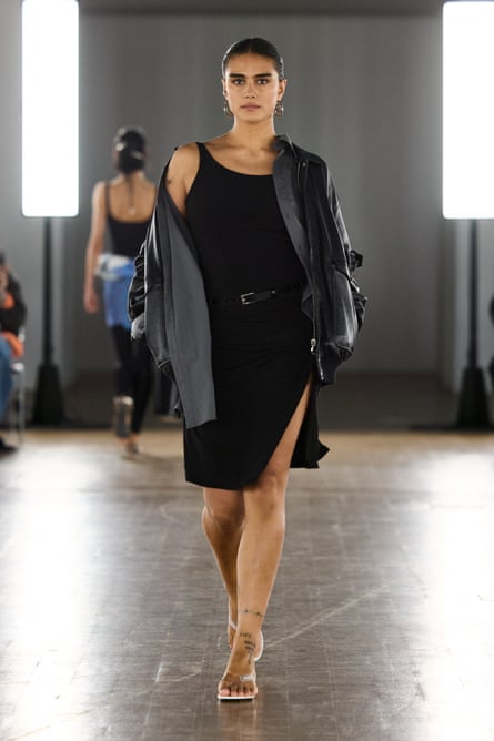 she wears a short black sleeveless dress with belt and a split up one thigh, and high-heeled sandals; she has a black jacket over her shoulders and her dark hair slicked back.