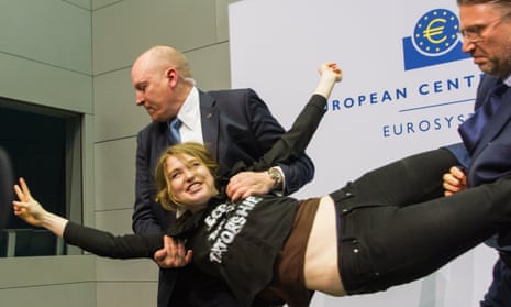 A protester who jumped on top of ECB president Mario Draghi’s desk during a news conference at the European Central Bank is detained by security. Her shirt reads “End the ECB Dick-tatorship”.