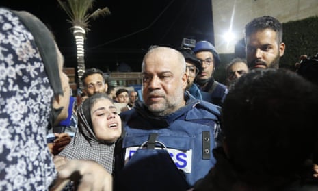 A middle-aged Palestinian man with a bald head, light white bread, blue flak jacket, looking stunned and standing in a crowd of people, with a young woman, her hair in a scarf, chin raised and crying beside him.