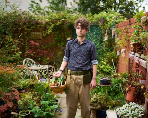 Samuel Rowe: he is 35 with dark wavy hair and stands holding a trug filled with vegetables in a pretty garden; there are pots of flowers on shelving on the brick wall, a flowerbed by paving stones, and antique-style table and chairs. He wears light tan trousers with a tool belt plus patterned dark blue shirt.