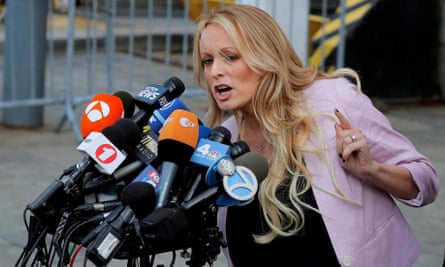 A white woman with long blond hair, a light pink blazer, and a black top holds up one finger in a gesture as she leans over and speaks into about a dozen microphones that are clustered together.