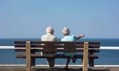 Elderly couple sitting on bench overlooking sea, UK.