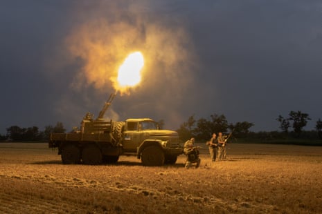 flames shoot from long guns mounted on a truck and more soldiers fire guns from the ground; they are in what appears to be a recently harvested cornfield.