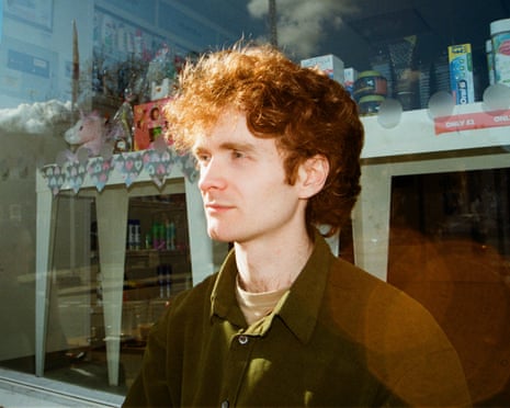 A ginger man wearing a brown jacket looks into the distance as he sits in front of a shop.