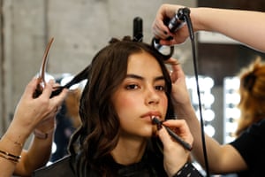 A model sits surrounded by pairs of hands that are applying make-up and styling her hair