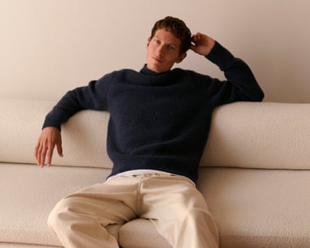 A man, wearing a navy sweater and cream trousers, reclines on a cream sofa in a cream room