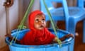 A baby wearing a red hijab sits in a blue bucket to be weighed