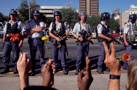 A row of white police officers wearing helmets and carrying automatic weapons faces what we see only as Black and brown hands raised holding flowers and showing the peace sign.