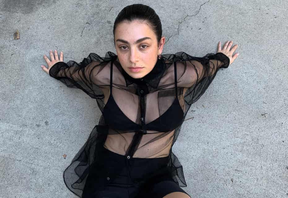Portait of singer and songwriter Charli XCX wearing a sheer black shirt, waist up, against grey stone background
