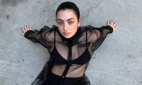 Portait of singer and songwriter Charli XCX wearing a sheer black shirt, waist up, against grey stone background