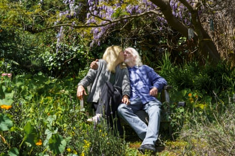 Gray-haired man and woman sit in shade under tree on green overgrown lawn, kissing.