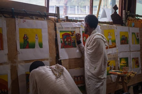 Two man wearing white robes work on series of religious paintings attached to boards along a wall