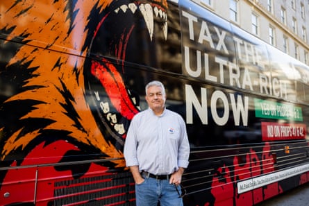 a man stands in front of a bus emblazoned with âÂÂtax the ultra-rich nowâÂÂ