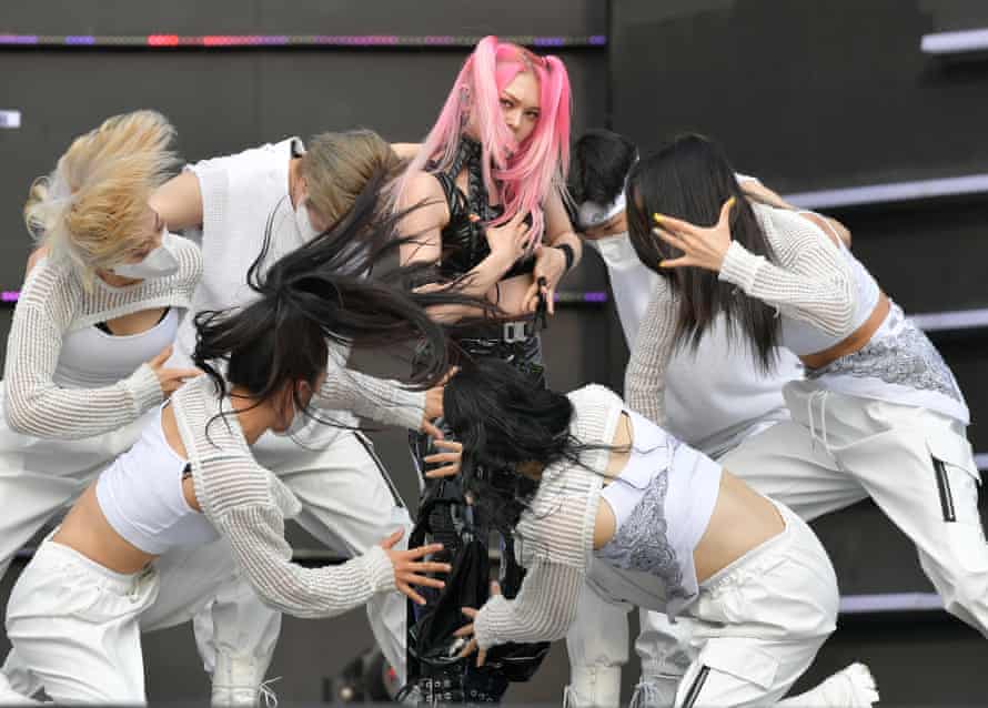 A woman dressed in black with pink hair performs on stage surrounded by dancers dressed in white