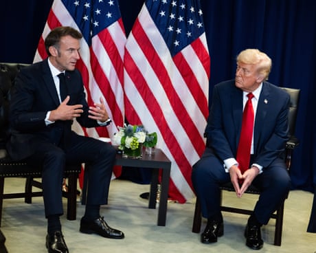 Trump and Macron sit on low chairs in front of two US flags; Macron is speaking while Trump looks on.