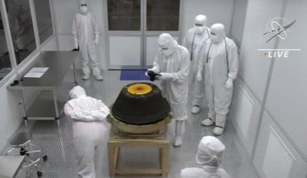 People in white full-body suits stand around a black and yellow circular object.