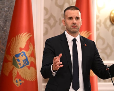 Milojko Spajić speaks in front of a red and gold heraldic flag and gold-patterned wallpaper in a ceremonial room. He has very short, neat dark hair and wears a black suit and tie. He is in his late 30s.