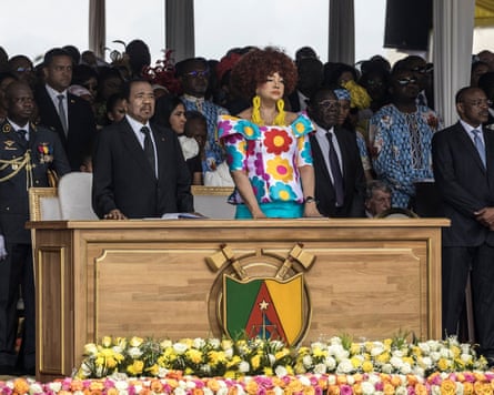 Paul Biya and Chantal Biya stand behind a wooden desk placed on a stage, as other people stand behind