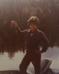In a grainy image, he is standing in a boat on a lake, with a forest in the background. He is holding what appears to be a fishing lure