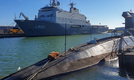 A wide shot of a walrus lying on the bow of a submarine foregrounded against a large naval ship.