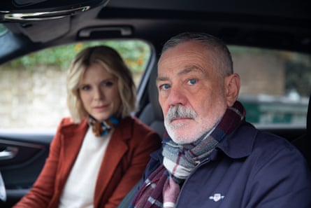 Emilia Fox in red jacket and a David Wilson in blue with plaid scarf sitting in a car and looking toward camera