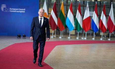 Olaf Scholz walks on a red carpet, with flags of EU countries in the background 
