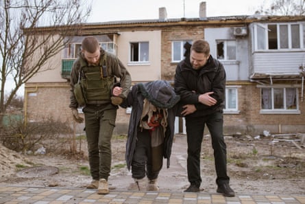Two men help a stooping woman outside a damaged house