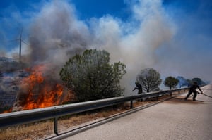Two people use a fire hose over the side of road as smoke fills the air from blaze burning throw trees and bushes