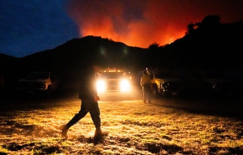 A firefighter walks past engines at night as wildfire glows on distant hills in Big Sur